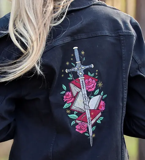 Embroidery on Jackets - image features a black jean jacket with a machine embroidery design of a sword with books